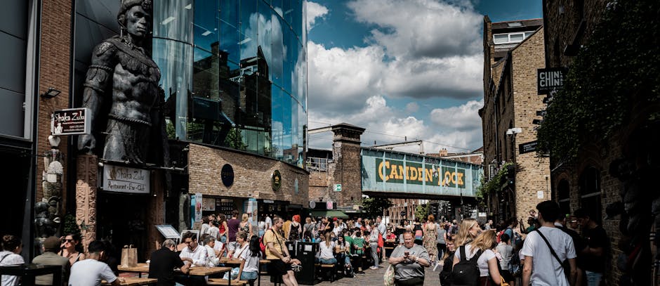 A busy outdoor scene in Camden Town showing the exterior of a modern glass-fronted building housing shops and eateries, with a large sign reading 'Camden Lock' hanging overhead. The area is crowded with numerous pedestrians, some seated at outdoor tables enjoying food and drinks, while others walk or stand around. Moving equipment such as cardboard boxes, plastic wrapping, and protective blankets are visible near the pavement, indicating an ongoing home relocation process. A man with a van is likely involved in the furniture transport, as part of a professional removals service. The environment is illuminated by natural daylight, with a partly cloudy sky overhead, and various vehicles and shops lining the street, reflecting the vibrant, bustling atmosphere of Camden Market and the logistical activities associated with packing and moving services like those provided by Man with Van Camden Town.