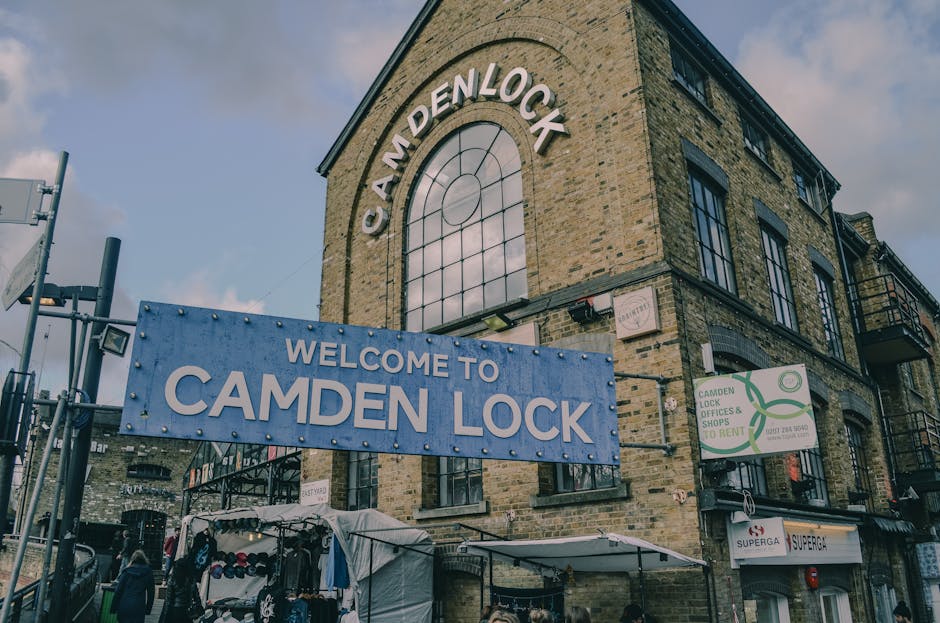 A busy outdoor scene in Camden Town showing the exterior of a modern glass-fronted building housing shops and eateries, with a large sign reading 'Camden Lock' hanging overhead. The area is crowded with numerous pedestrians, some seated at outdoor tables enjoying food and drinks, while others walk or stand around. Moving equipment such as cardboard boxes, plastic wrapping, and protective blankets are visible near the pavement, indicating an ongoing home relocation process. A man with a van is likely involved in the furniture transport, as part of a professional removals service. The environment is illuminated by natural daylight, with a partly cloudy sky overhead, and various vehicles and shops lining the street, reflecting the vibrant, bustling atmosphere of Camden Market and the logistical activities associated with packing and moving services like those provided by Man with Van Camden Town.