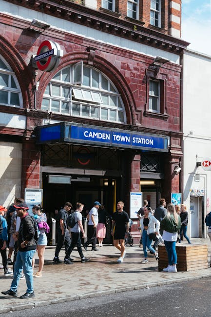 Exterior view of Camden Town underground station in London, showing a red-brick façade with large arched windows and a blue sign reading 'CAMDEN TOWN STATION' above the entrance. Several pedestrians, some carrying backpacks and shopping bags, are walking across the pavement in front of the station, with a mix of men and women dressed in casual attire. There is a wooden bench on the sidewalk and a red roundel tube station sign mounted above the entrance. The scene captures a busy urban environment typical of home relocation and moving logistics near a London transport hub, with natural daylight illuminating the area.