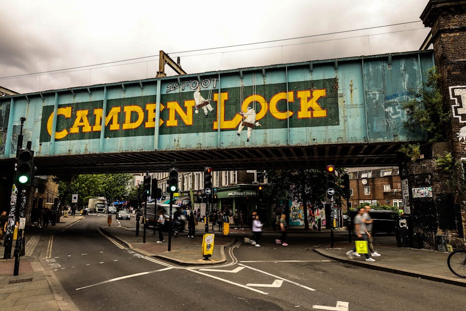 A street scene in Camden Town featuring a large, weathered railway bridge overhead with the name 'CAMDEN LOCK' painted in bold yellow letters on a blue-green background. The bridge is equipped with skateboarding illustrations and has a mix of rust and chipped paint visible, suggesting age. Below, the busy street is filled with pedestrians, some walking and others waiting at crosswalks, with traffic lights, bollards, and road markings. Several people are carrying or handling cardboard boxes, plastic-wrapped furniture, and other packing materials as part of home relocation or furniture transport activities managed by Man with Van Camden Town. A van is parked nearby, likely involved in the moving process, and the environment features typical urban surroundings with buildings, trees, and street signage. The weather appears cloudy, and the scene captures the logistics and movement involved in packing, loading, and transporting household items during a house removal in an urban setting.